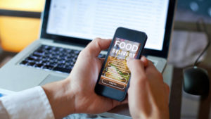 A pair of hands are holding a cell phone with a food delivery app open on the screen - an open laptop is in the background.